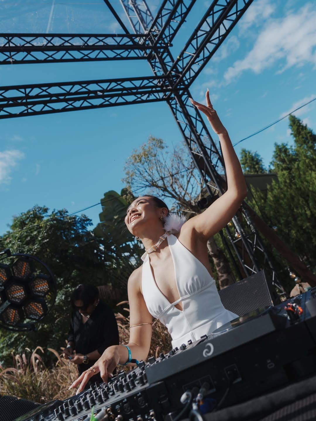 DJ in white with one arm raised behind the decks, blue sky and truss above